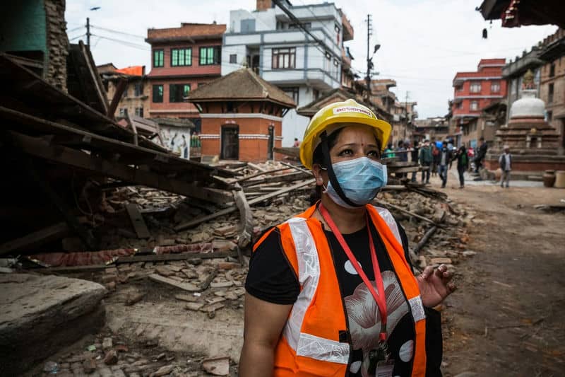Safety worker with mask and helmet inspects earthquake damage in urban area; debris from collapsed buildings visible.
