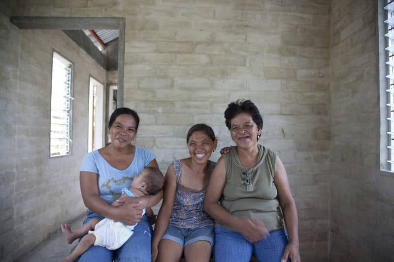 Three women sitting together, smiling inside a room with brick walls, one holding a baby.