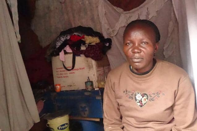 A person sitting in a modest room with clothes and household items in the background.