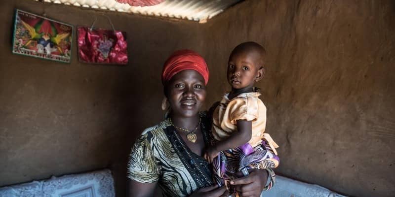 Mother holding child in modest home, conveying warmth and resilience. Simple decor with vibrant headscarf visible.