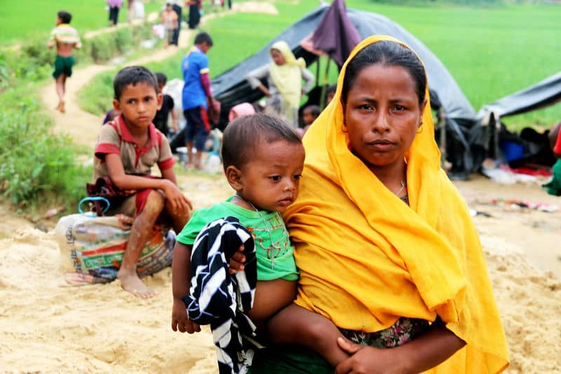 Mother holding child with others in background at a camp setting, amidst greenery and tents, depicting a refugee scene.
