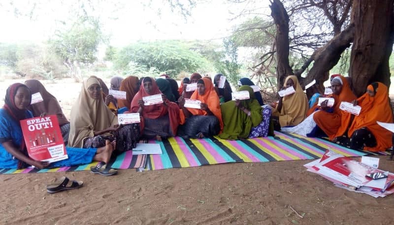 A group of women gathered outdoors, holding signs advocating for social change, seated on colorful mats under a tree.