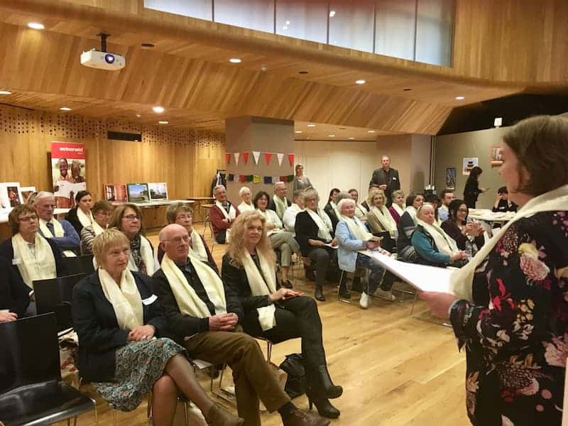 Audience listens to a speaker at an indoor event; participants wearing white scarves in a wood-paneled room.