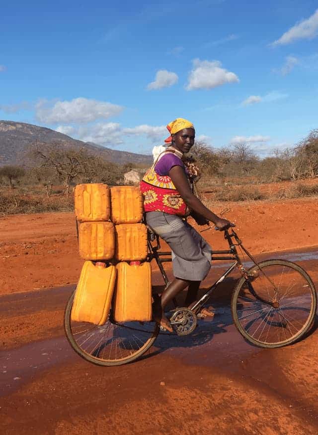 Person on a bike carrying yellow containers in rural landscape under blue sky.