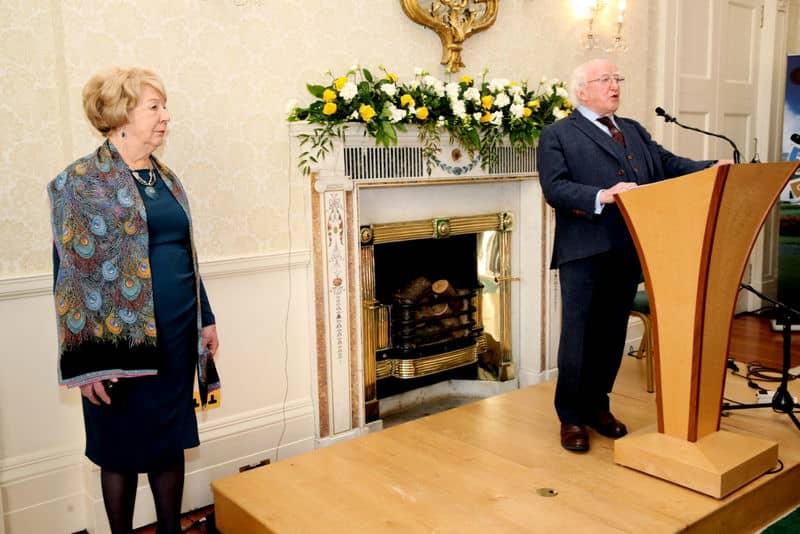 Elderly speaker at podium in elegant room with floral decor, attended by woman in a scarf, near a classic fireplace.