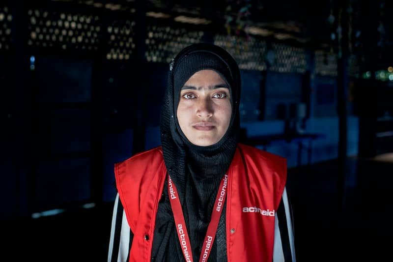 Volunteer wearing an ActionAid vest stands in a dimly lit room, conveying dedication and resilience.