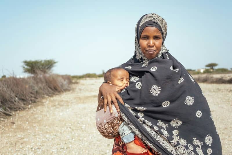 Mother holding sleeping baby outdoors in traditional attire, standing on a rural pathway under clear blue sky.