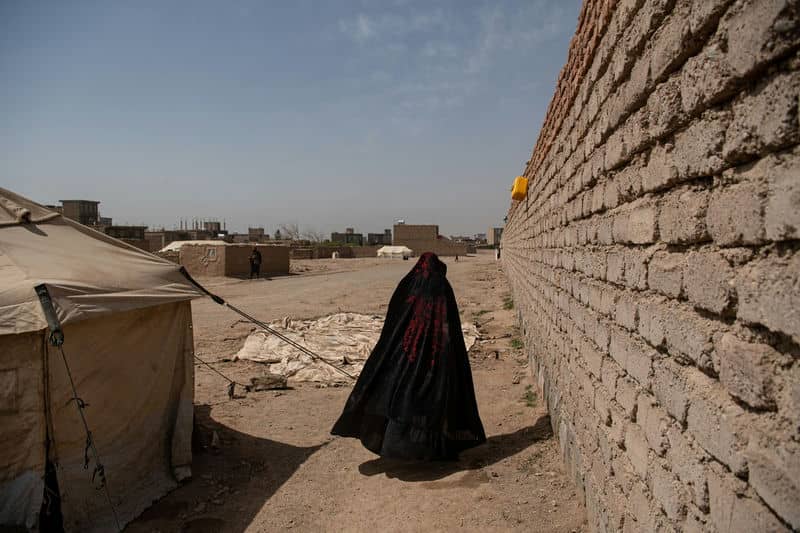 Woman in black cloak walks between brick wall and tent in a dusty settlement under a clear blue sky.