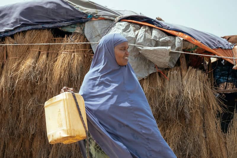 Woman carrying a yellow container in front of a thatched-roof shelter, wearing a blue hijab.