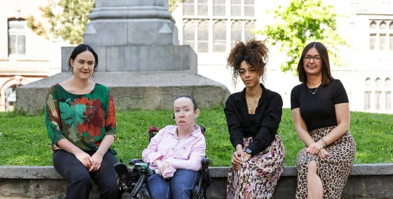 Four women sitting on a park bench, smiling, with a historical building in the background on a sunny day.