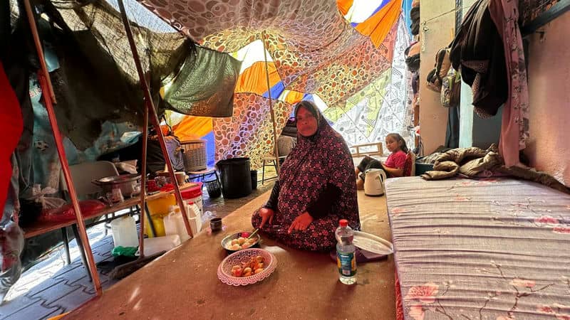 Woman sitting in colorful tent with food plates, child in background, cozy interior setting with various objects.