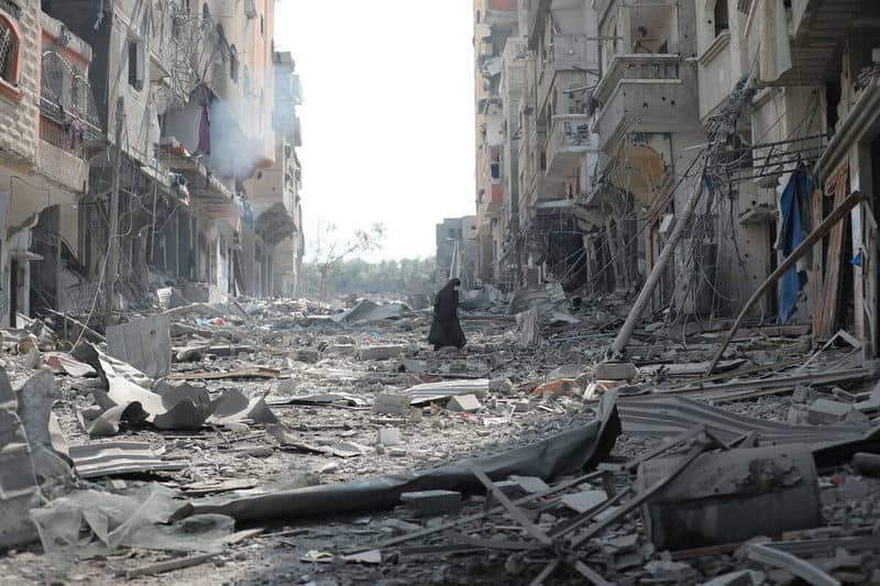War-torn street with debris and damaged buildings, lone figure walking amid destruction.