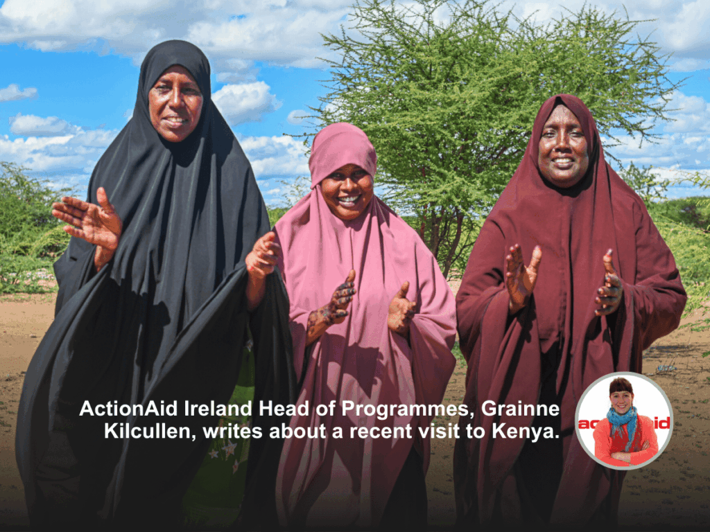 Three women smiling in colorful headscarves, outdoors in Kenya under a blue sky with green foliage in the background.
