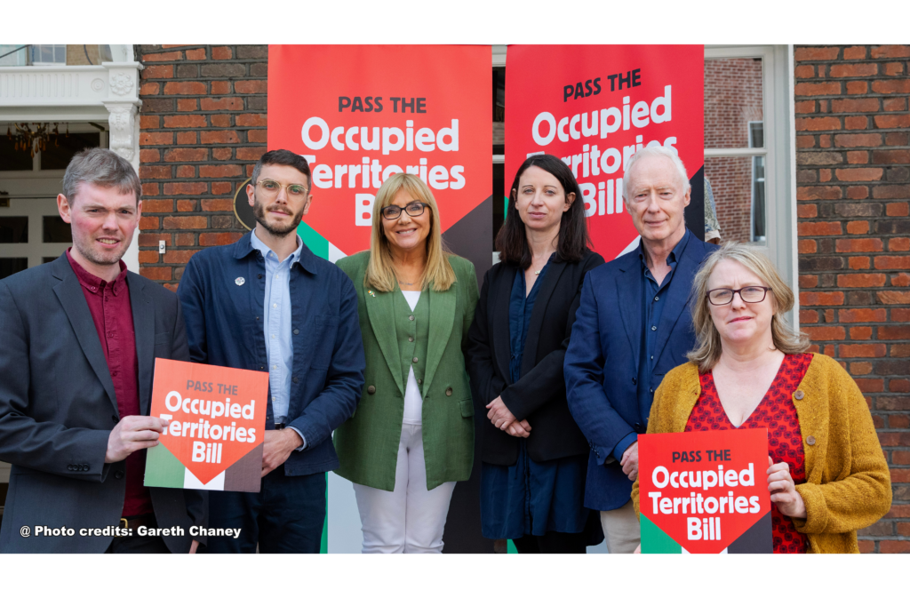 Group supporting the Occupied Territories Bill, holding signs and standing in front of banners in an outdoor setting.