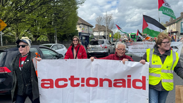 Protesters carry an ActionAid banner during a street march with flags in the background, highlighting activism.