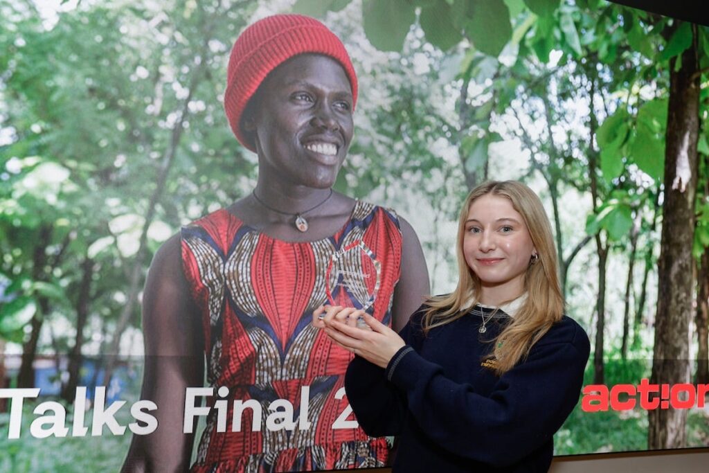Young woman holding an award stands smiling in front of a joyful, nature-themed poster with greenery background.