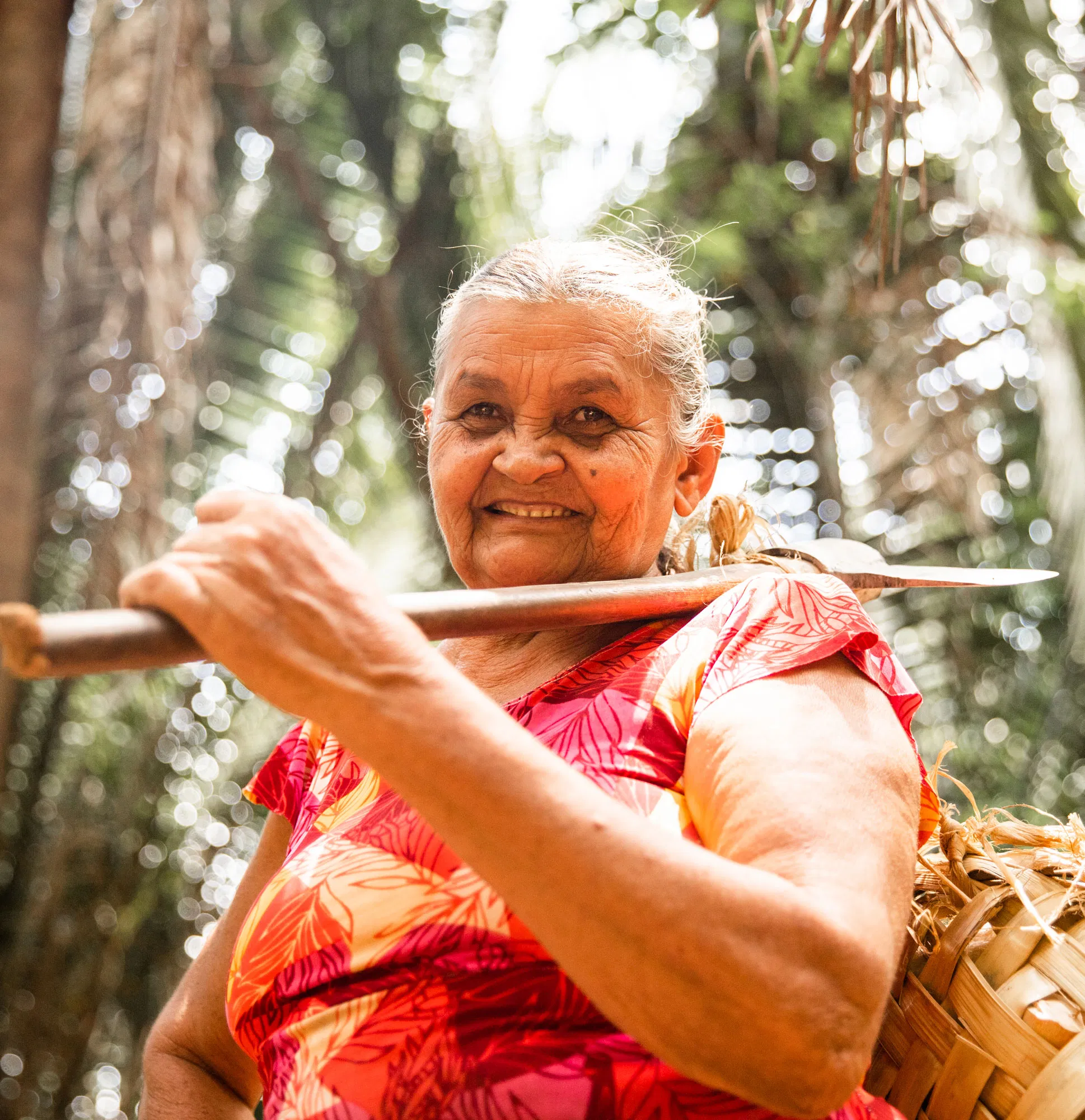 Elderly woman in vibrant clothing smiles while holding a tool in a lush, sunlit forest setting.