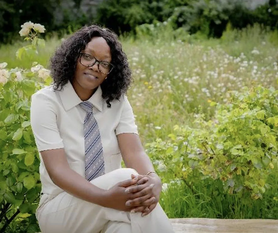 Person in white attire and tie seated in a garden, surrounded by lush greenery and flowers.