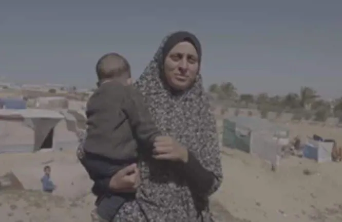A woman holds a child outside in a refugee camp setting, against a backdrop of makeshift shelters.