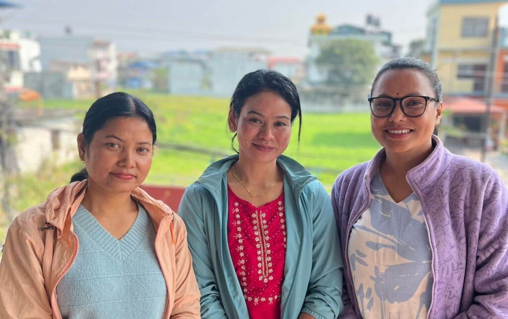 Three women smiling outdoors with a blurred green and urban background, wearing casual jackets and shirts.