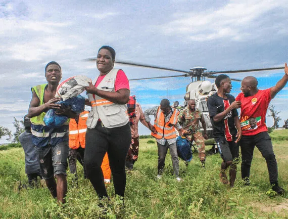 Rescue workers and volunteers deliver supplies via helicopter in a grassy field under cloudy skies.
