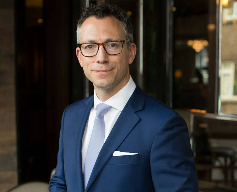 Smiling man in a blue suit with glasses, standing indoors, professional business setting.