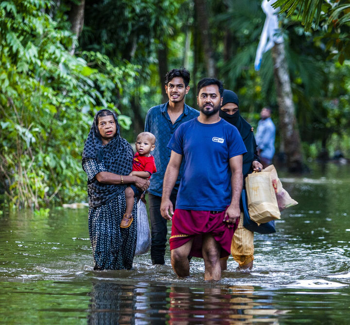 Family wading through floodwaters in tropical setting, carrying supplies and a child, surrounded by lush greenery.