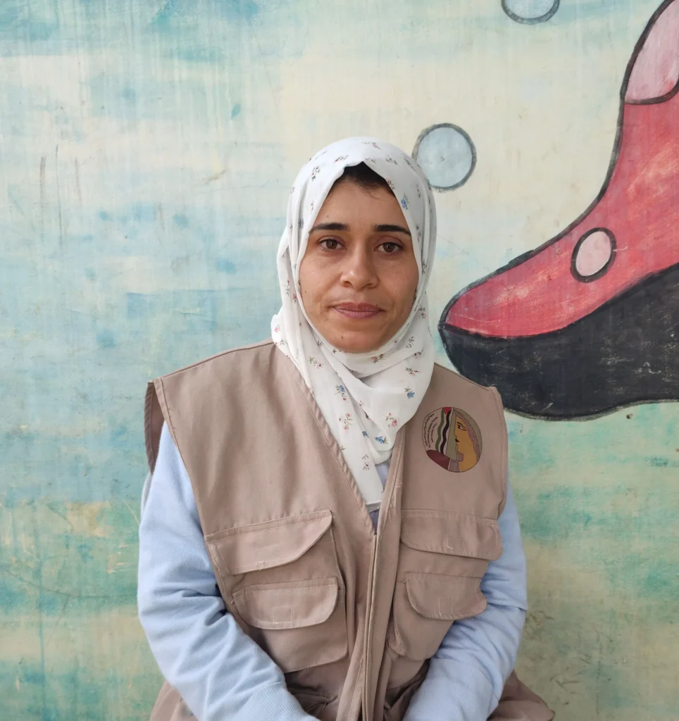 Woman in a hijab and vest outside, standing by a mural with a red boot illustration.