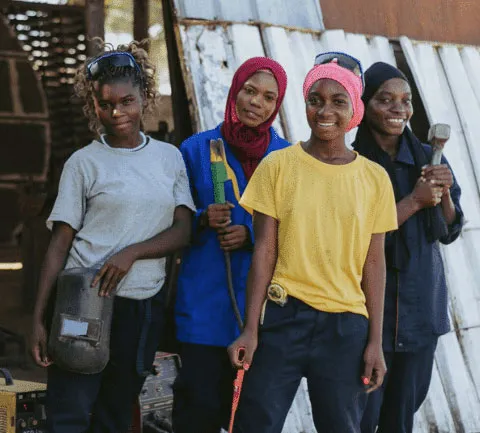 Women in construction gear smiling, holding tools, standing together outside a workshop. Empowerment and teamwork in trades.