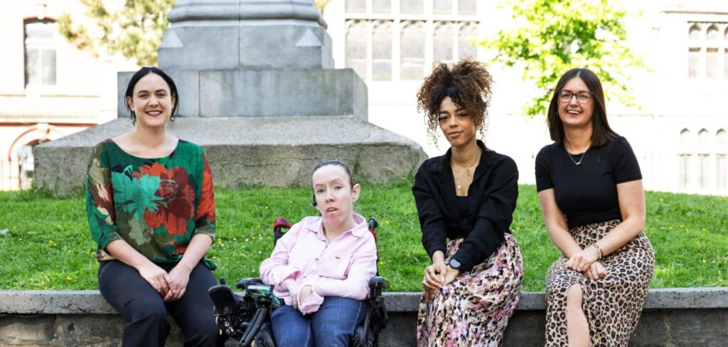 Four diverse women sitting on a stone ledge in a park on a sunny day, smiling at the camera.