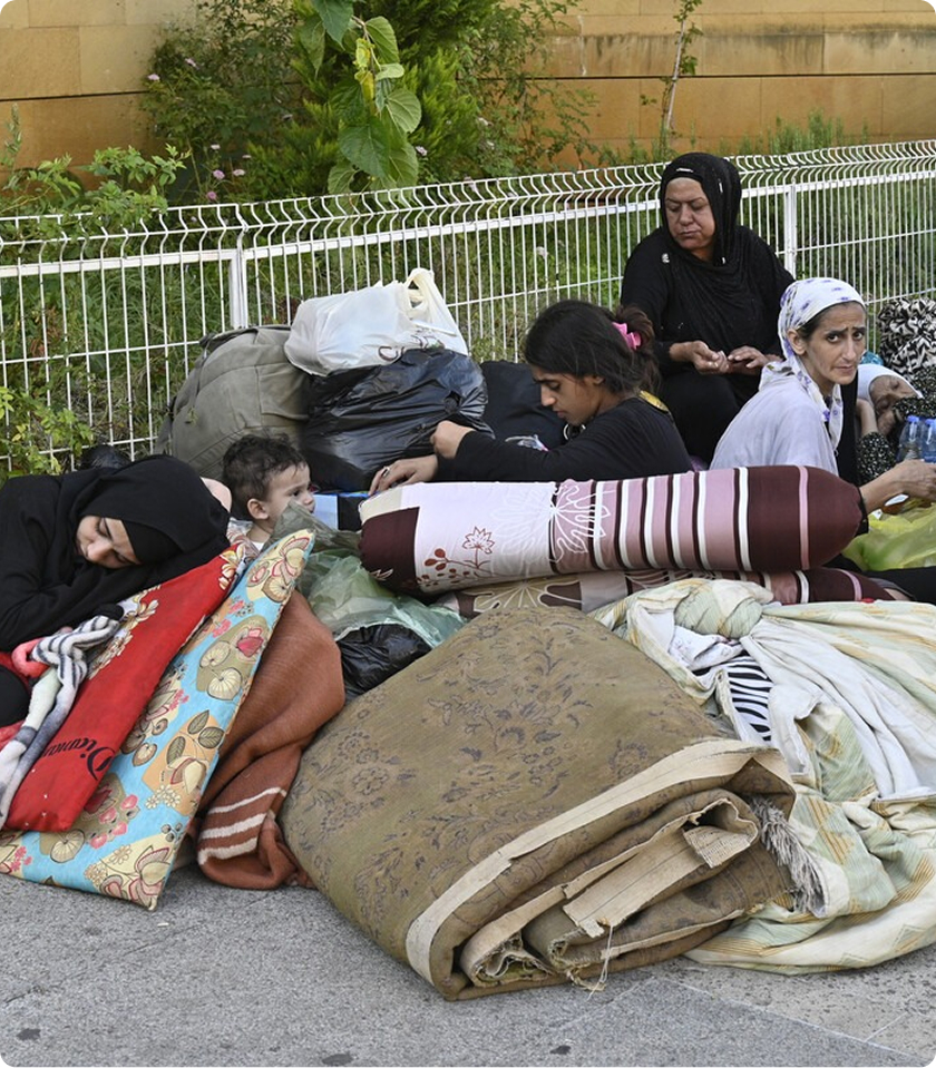 People resting on folded blankets and pillows outdoors near a fence, surrounded by bags and greenery.