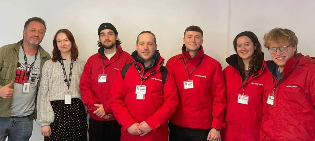 Team in red jackets with ID badges posing for a group photo against a plain background.