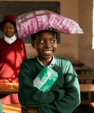 Smiling student in classroom balancing sanitary napkins on head, holding a pack, promoting menstrual hygiene awareness.