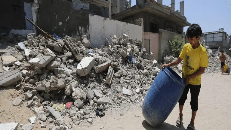 Child moving a blue barrel near rubble in a conflict-affected area, highlighting post-disaster recovery efforts.